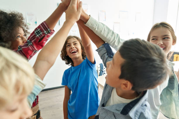 Happy diverse kids school students group giving high five together in classroom. Happy diverse multiethnic kids junior school students group giving high five together in classroom. Excited children celebrating achievements, teamwork, diversity and friendship with highfive concept. mixed racial group of friendly children laughing together stock pictures, royalty-free photos & images