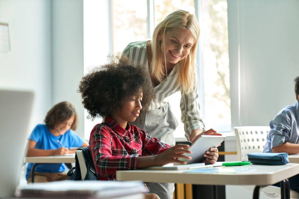 maestra que ayuda a una colegiala africana que usa tabletas en clase en el aula. - education school computer teacher fotografías e imágenes de stock