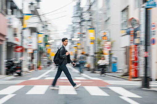 Blurred motion side view of man in early 20s wearing jeans, black leather jacket, and carrying backpack while walking in zebra crossing.