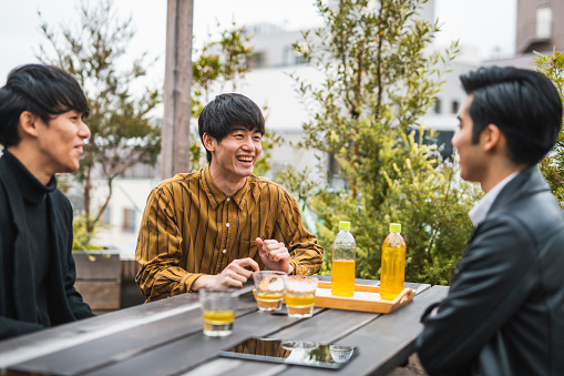 Men in early 20s sitting at picnic table on Tokyo rooftop laughing as they enjoy bottled tea and conversation.