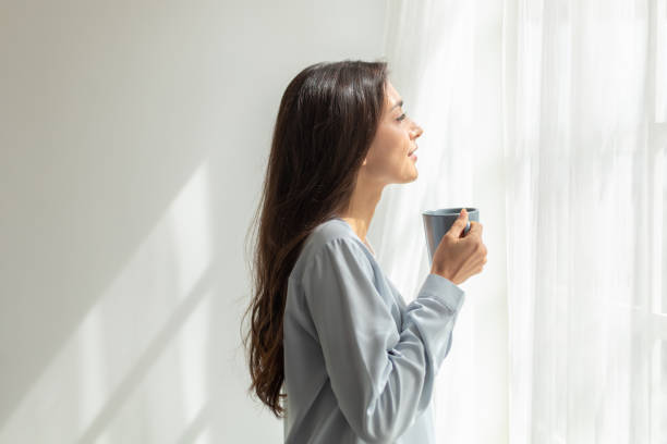 Happy woman stand drink coffee and opening window curtains breathe fresh air stretch exercise in bedroom. Female overjoyed welcome new life at new home. Optimism and mental health concept stock photo