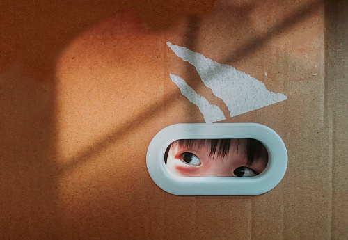 asian chinese baby boy peeking through ellipse hole on cardboard box hiding in cardboard box, looking out with window frame shadow