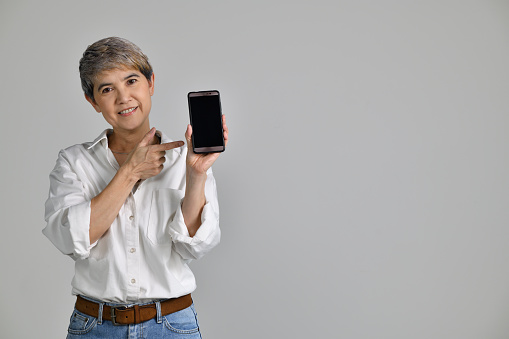 Middle aged Asian woman showing blank smartphone screen and pointing on it while looking at the camera isolated on white background