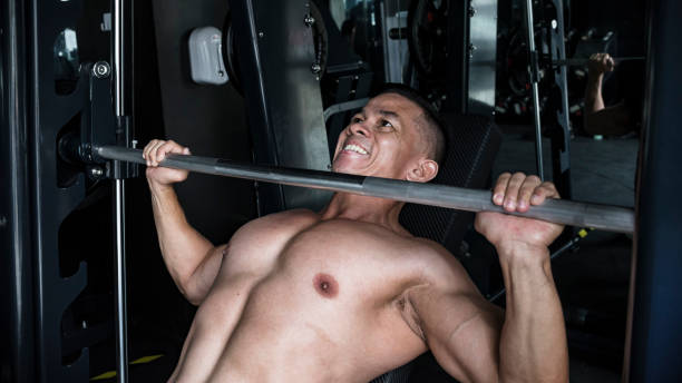 A bare chested man in the middle of doing intense incline barbell bench presses on a Smith Machine at a gym or fitness center. In the zone and motivated doing chest muscle training.