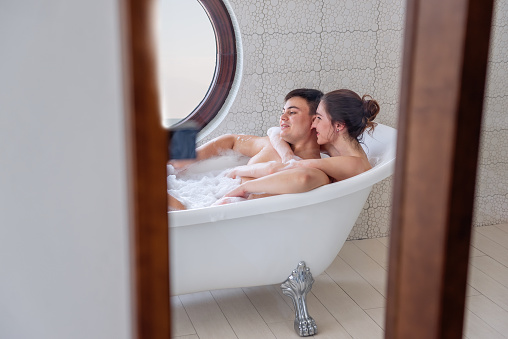 A loving couple takes a bubble bath in a minimalist white, modern interior. A young girl hugs a man from behind, lovers kiss, laugh. In the morning of the Active Lux Weekend. Journey of the newlyweds