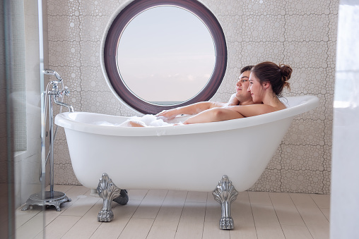 A loving couple takes a bubble bath in a minimalist white, modern interior. A young girl hugs a man from behind, lovers kiss, laugh. In the morning of the Active Lux Weekend. Journey of the newlyweds