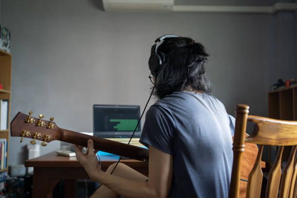 A man with long hair wearing headphones and playing acoustic guitar A man with long hair wearing headphones and playing acoustic guitar in order to recording into a computer in a domestic room making music stock pictures, royalty-free photos & images