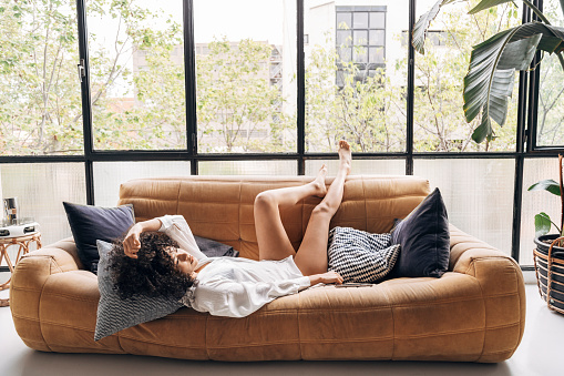 Beautiful young african american woman lying down on sofa with legs up in a bright loft living room. Home concept.