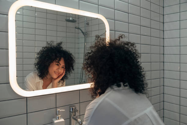 Young mixed race woman looking herself in the mirror with love. Smiling female with curly hair in a modern washroom. Young mixed race woman looking herself in the mirror with love. Smiling female with curly hair in a modern washroom. Self love concept. looking in mirror stock pictures, royalty-free photos & images