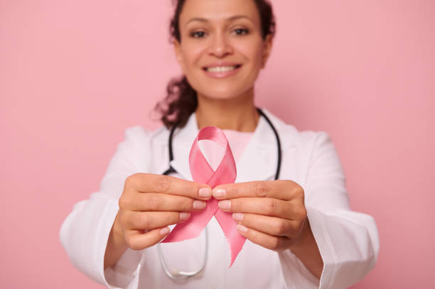 foco suave na fita rosa de conscientização do câncer de mama, nas mãos de médica mestiça desfocada em jaleco médico branco, isolada em fundo colorido. 1º de outubro, dia mundial de combate ao câncer de mama - breast cancer awareness ribbon breast cancer ribbon charity and relief work - fotografias e filmes do acervo