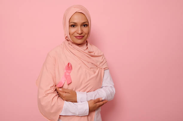 mulher muçulmana deslumbrante com a cabeça coberta de hijab, fica em fundo rosa com braços cruzados no peito e usando uma fita rosa, símbolo do dia da conscientização do câncer de mama. conceito médico, espaço de cópia - breast cancer awareness ribbon breast cancer ribbon charity and relief work - fotografias e filmes do acervo