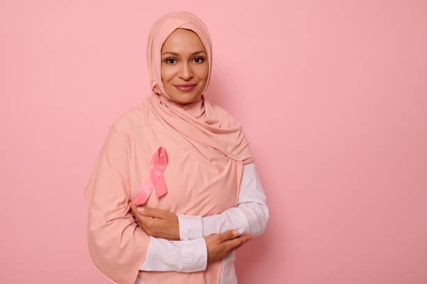 великолепная мусульманка с покрытой головой в хиджабе, стоит на розовом фоне со скрещенными руками на груди и носит розо�вую ленту, символ д� - cancer breast cancer breast cancer awareness ribbon charity and relief work стоковые фото и изображения
