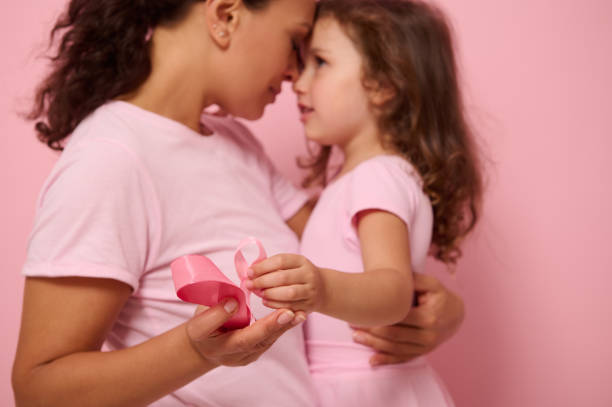 foco em fitas de cetim rosa, símbolo do dia internacional de conscientização do câncer de mama, nas mãos da mãe amorosa desfocada e do fundo da filha adorável. conceito de saúde feminina e educação médica - breast cancer awareness ribbon breast cancer ribbon charity and relief work - fotografias e filmes do acervo