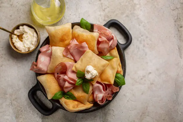 Italian  appetizer. Fried bread crescentine or gnocco fritto with mortadella and soft cheese, decorated with basil leaves. Light stone background.