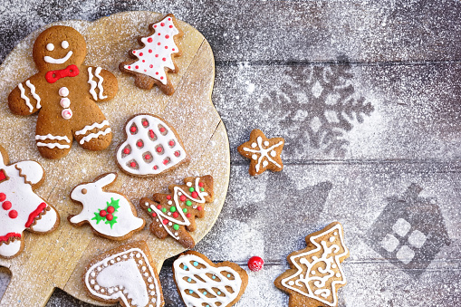 Christmas cookies of various shapes in sugar glaze on a cutting board on a brown wooden table sprinkled with flour, flat lay, copy space. Christmas composition with gingerbread men, fir-trees, rabbit.