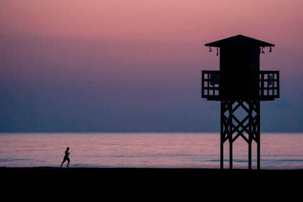 海を背景にしたライフガード小屋のバックライトとビーチ沿いを走る人のシルエット - lifeguard orange nature beach ストックフォトと画像