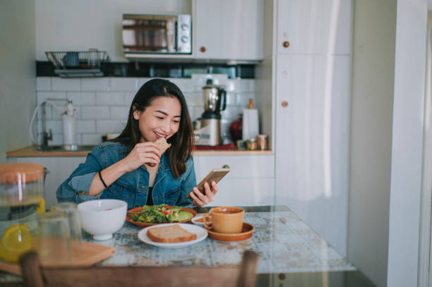 bella donna cinese asiatica che legge sul suo smartphone e si gode la colazione a casa imburre il burro di arachidi di pane integrale con caffè - non alcoholic beverage comfort food snack dining table foto e immagini stock