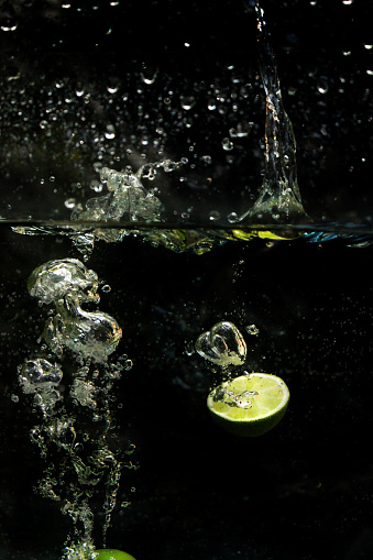 Stock photo showing close-up view of water droplets and bubbles from dropping pieces of sliced lime fruit into water, pictured against a black background.