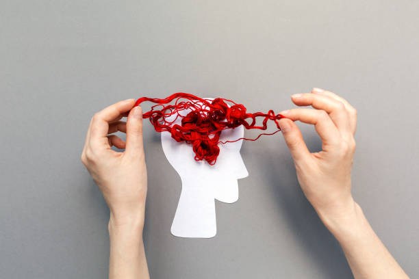 女性の手は、頭のシルエットに絡まった赤い糸を解き明かし、脳を表します。灰色の背景。フラットレイ。メンタルヘルスと精神学問題の概念 - 精神障害 ストックフォトと画像