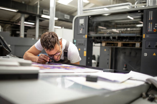 Professional print worker checking imprint quality and color matching with magnifying glass in printing house. Professional print worker checking imprint quality and color matching with magnifying glass in printing house. printing plate photos stock pictures, royalty-free photos & images