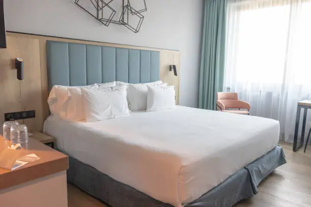 Light and cozy modern hotel room with comfortable king-size bed and many pillows, large window and blue headboard.