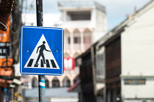 A pedestrian crosswalk signpost symbol in blue and triangle logo on the street with city as blurred background. Transportation sign object photo.