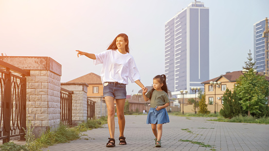 Young mother with flying hair walks with Korean little girl along contemporary city embankment pointing to beauty at sunset