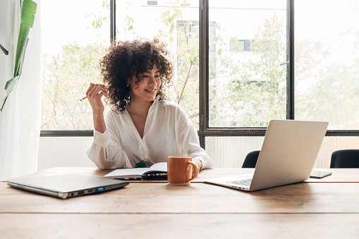 Smiling, happy, Young beautiful african american woman studying at home with laptop. Taking notes in notebook. Bright spacious living room. E-learning concept. Study at home concept.