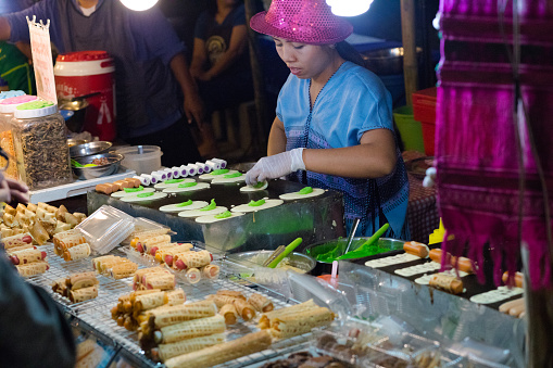 Blue dressed thai woman with pink hat is selling spring rolls on night market in Chiang Mai. Woman is also selling small sweet  thai pancakes. Woman is preparing those pancakes