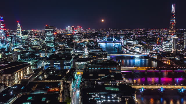 Fly along Thames river at night. Hyperlapse of cityscape with city lights and full Moon. The Shard, skyscrapers in City financial hub and Tower Bridge. London, UK