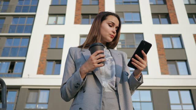 Young woman in a business suit looks at the phone and drinks coffee from a plastic cup on the background of a building