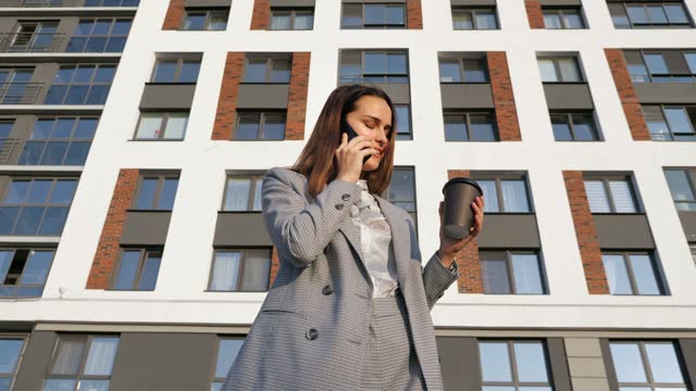 woman in a business suit drinks coffee from a plastic cup and talks on the phone against the background of a building on a sunny day
