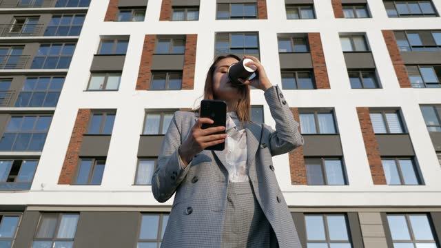 woman in a business suit drinks coffee from a plastic cup and looks into the phone against the background of a building