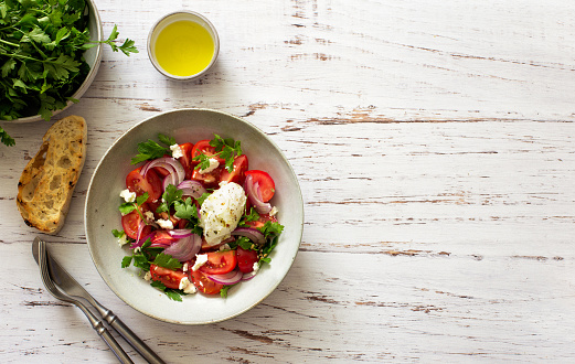 Tomatoes, red onions, greens and soft cheese salad in a bowl on white wooden background. Flat lay. Copy space
