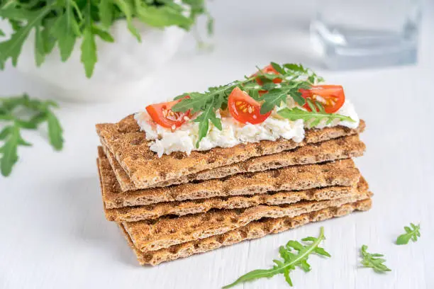 Pile of crispbread - a flat and dry type of healthy cracker, containing whole wheat flour with ricotta, cherry tomato slices and rocket leaf  topping on white wooden background served for breakfast