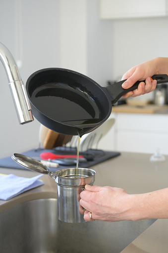 Pouring used cooking oil from an old frying pan into a metal cooking container in her kitchen.Oil recycling concept
