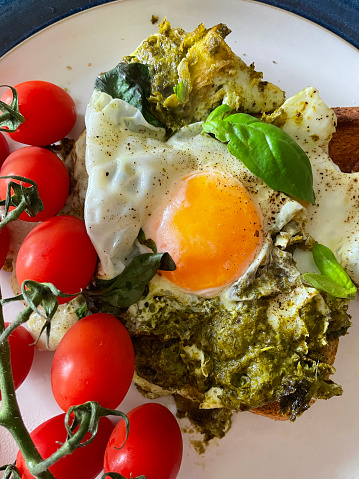 Stock photo showing an elevated view of blue rimmed plate containing a fried breakfast, with pesto, fried eggs sunny side up, cherry tomatoes and wholegrain toast garnished with basil leaves.