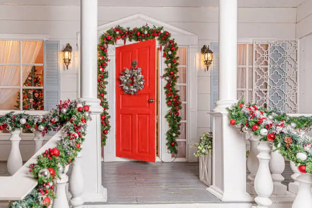 Photo of Christmas porch decoration idea. House entrance with red door decorated for holidays. Red and green wreath garland of fir tree branches and lights on railing. Christmas eve at home