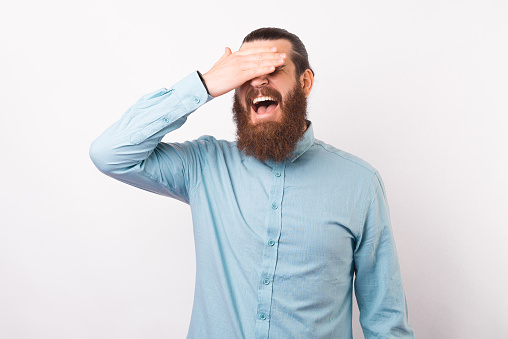 Bearded man wearing a shirt is making a facepalm gesture over white background.
