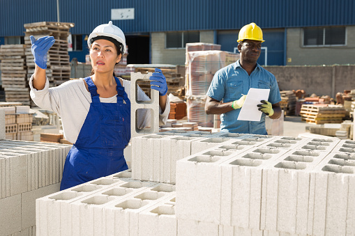 African-american man and Asian woman standing in outdoor construction material storage. Woman carrying hollow concrete block and calling out for someone.