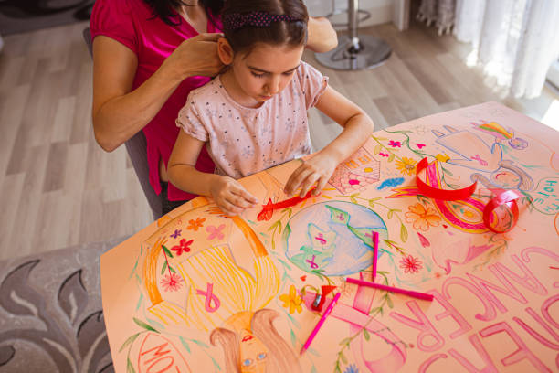 mutter macht ihre tochter frisur, während sie ihr hilft, ein poster während des brustkrebsmonats zu machen - community outreach connection child paper stock-fotos und bilder