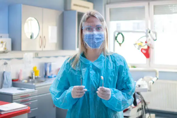 Female dentist in a protective suit with a protective shield and mask holding dental tools in dental office.