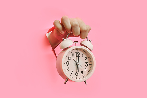 A hand holding an alarm clock though pink torn paper background