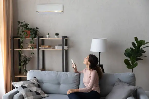 Young 30s Latina woman sitting on couch in living room holding remote controller turns on air conditioner set comfortable temperature inside, enjoy fresh air. Concept of climate control, modern house