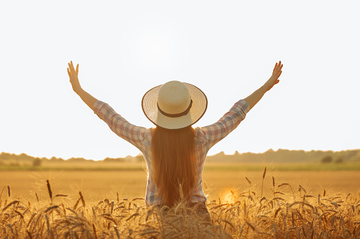 Rear view silhouette of woman with hat on farmer's head with hands raised up at sunset.