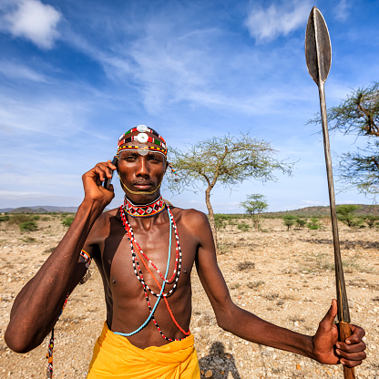 African warrior from Samburu tribe standing on savanna and using a mobile phone, central Kenya. Samburu tribe is one of the biggest tribes of north-central Kenya, and they are related to the Maasai.