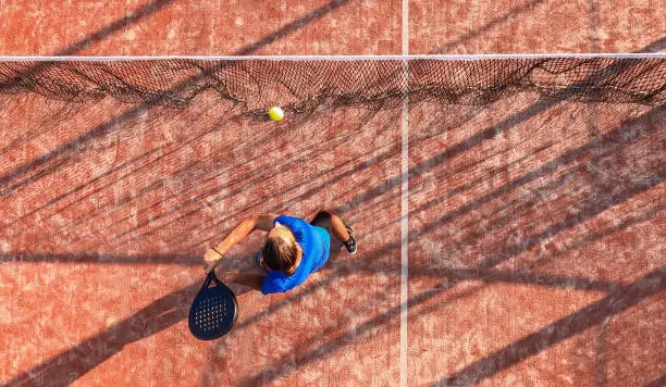 View from above of a professional paddle tennis player hitting the ball with the racket near the net of an outdoor court.