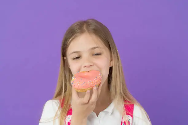 Child eat donut on violet background. Little girl bite glazed ring doughnut. Kid smiling with junk food. Sweet mood. Unhealthy eating and snack food.