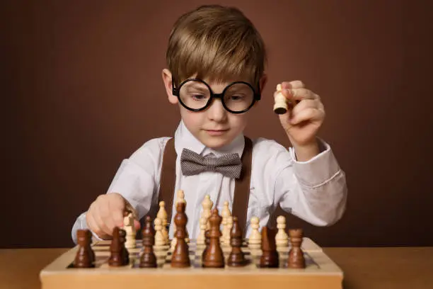 Little Kid playing Chess Game. Intelligent Small Cute Boy in Eyeglasses next to Chessboard holding Pawn. Child Education and Development Concept. Dark Brown Studio Background