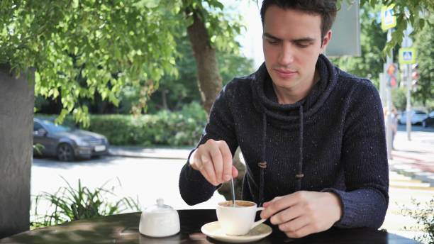 istock Young Man Mixing Sugar and Drinking Coffee, Sitting in Cafe Terrace 1338612081